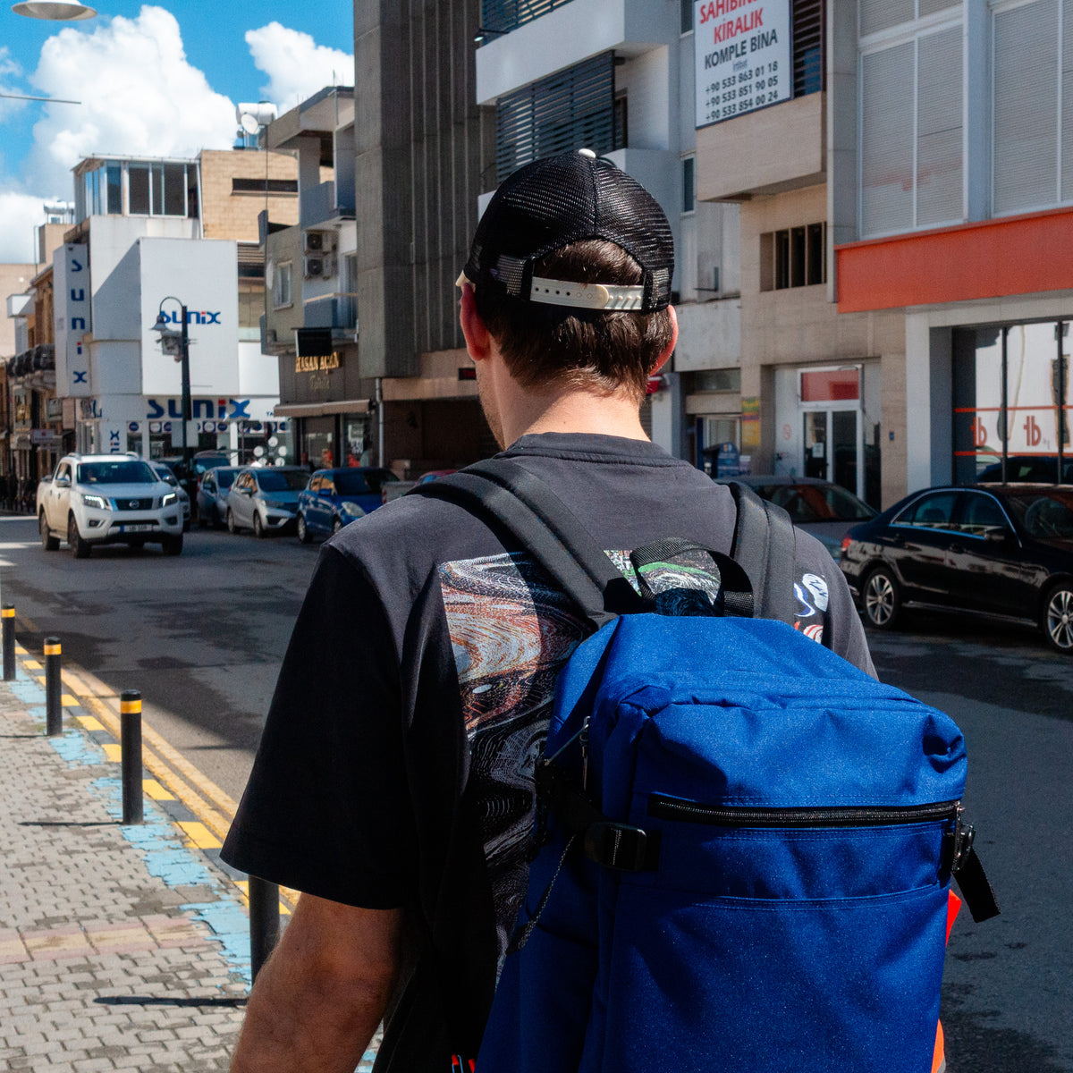 Person with ap and a blue backpack with vintage wash black t-shirt walking on a city street