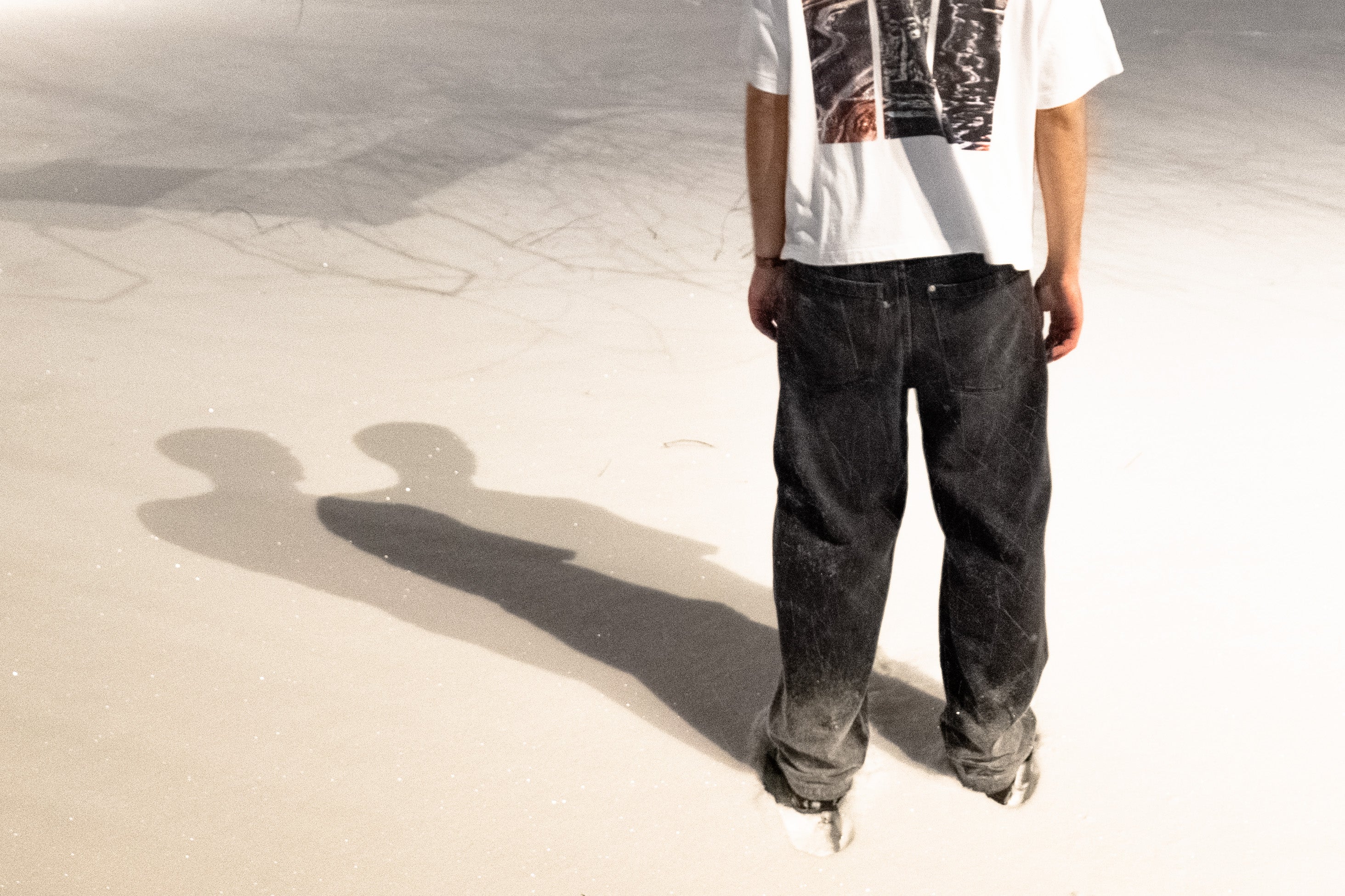 Man wearing a white t-shirt with DTG print and dark jeans standing on a snowy surface with a long shadow cast on his left.
