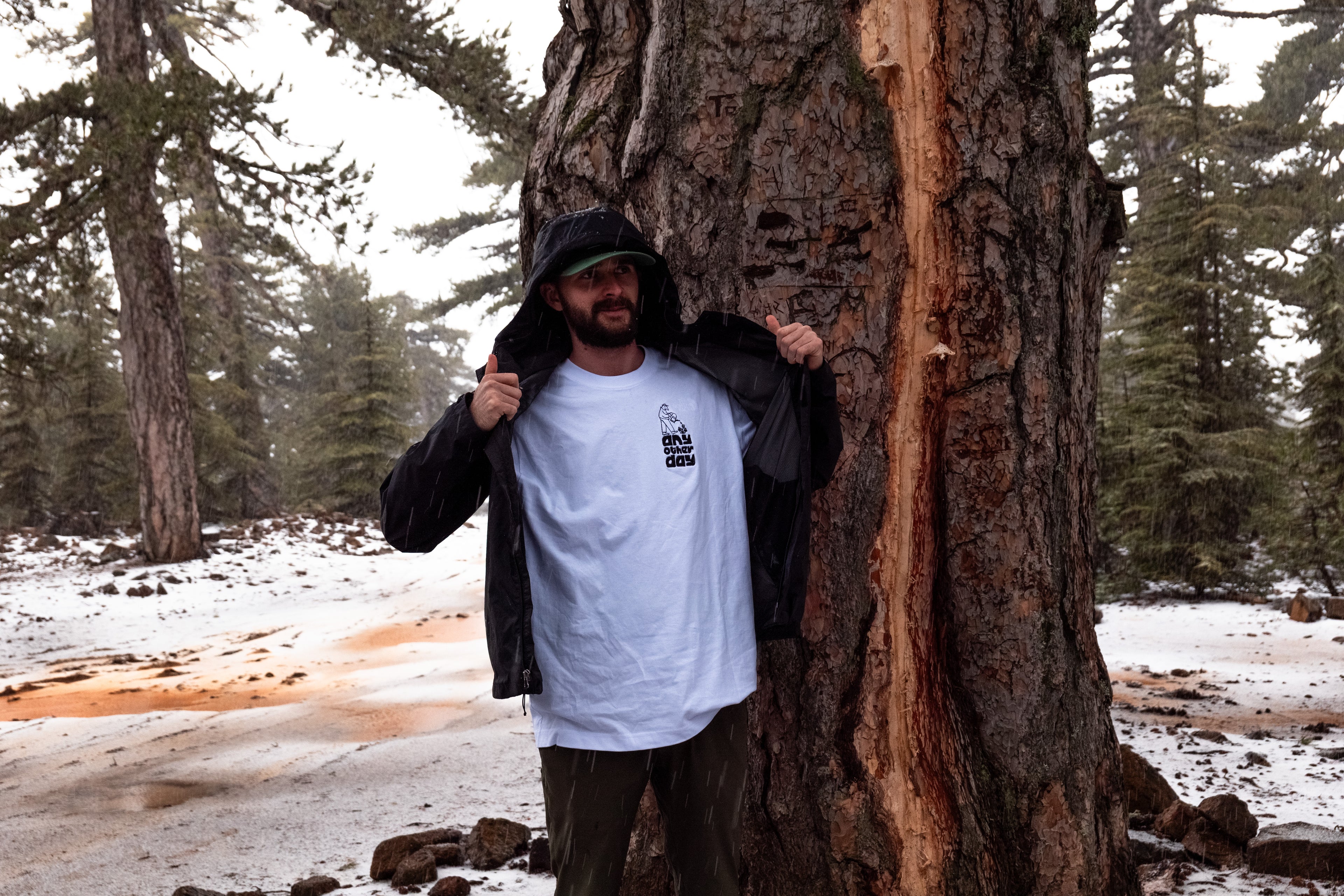 man with white embroidered t-shirt and jacket posing in front of a massive tree in a snowy forest
